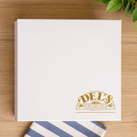 White box with 'DELS Popcorn Shop' logo on a wooden surface with a striped cloth and plant.