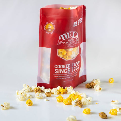 Red bag of Del's three kinds of Popcorn with popcorn scattered around on a white background